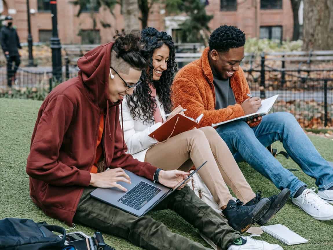 Three students sitting on the grass together, working on a laptop and notebooks